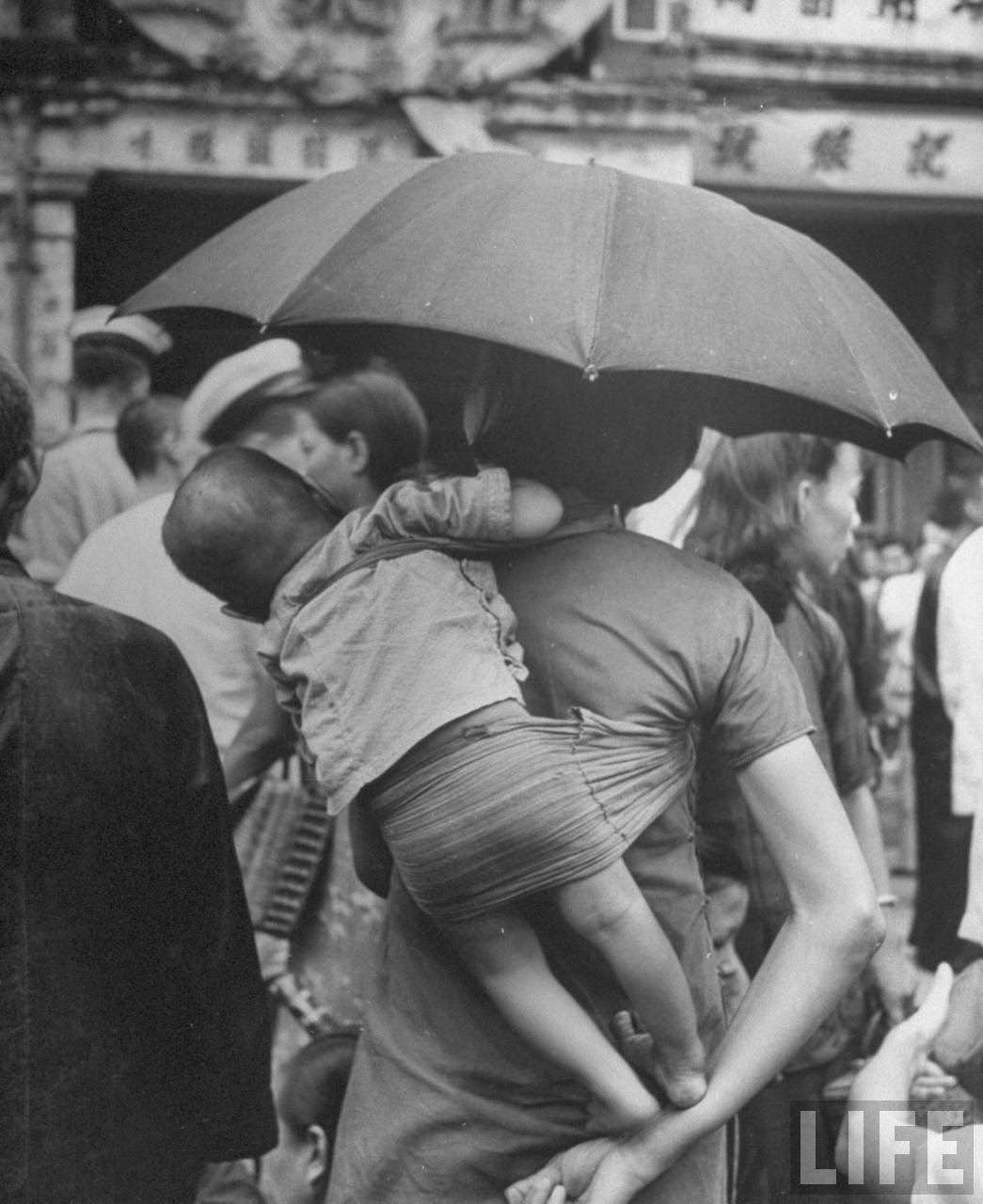 Woman carrying young child on her back through marketplace on Queen's Road.