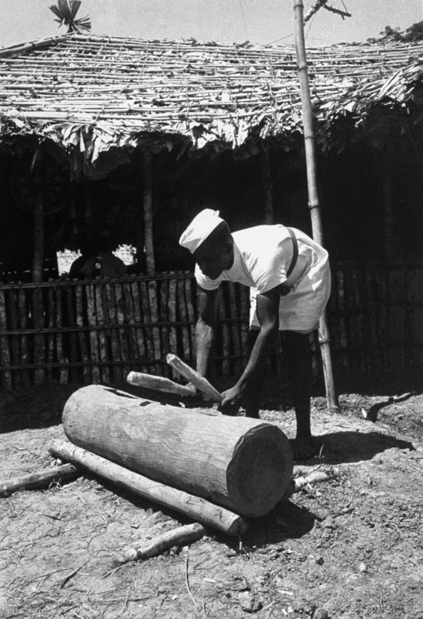 A man beats a hollow log with sticks — the daily 6 a.m. wake-up call for the cast and crew of The African Queen.