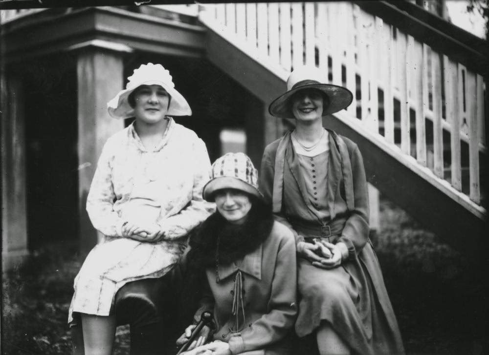 Three women wearing hats and knee-length dresses with long coats on a lounge chair, 1920s