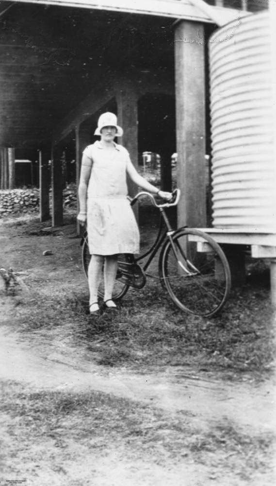 Woman wearing a knee-length skirt and hat posing next to a water tank, 1920s