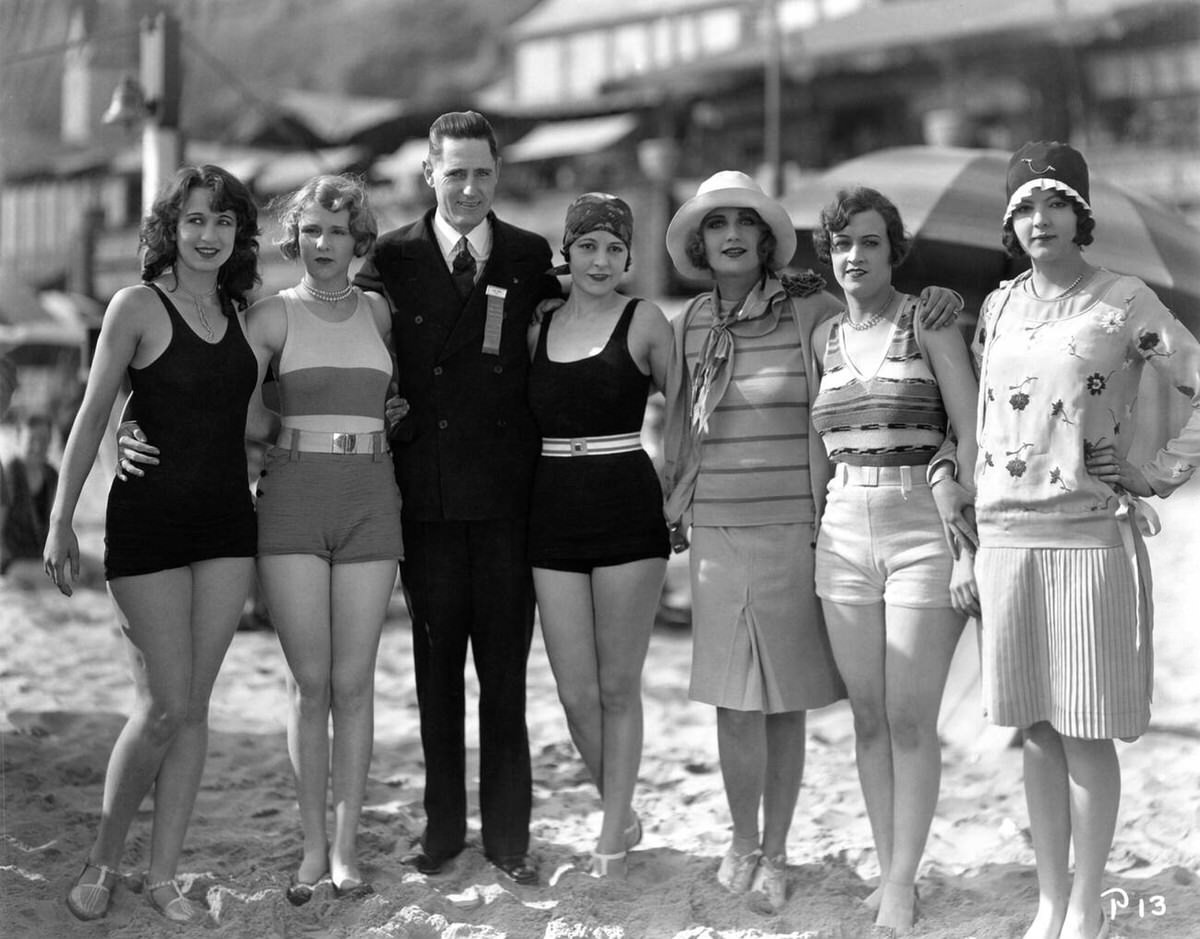 Mack Sennett Bathing Beauties posing with a movie theater manager on the beach, 1928
