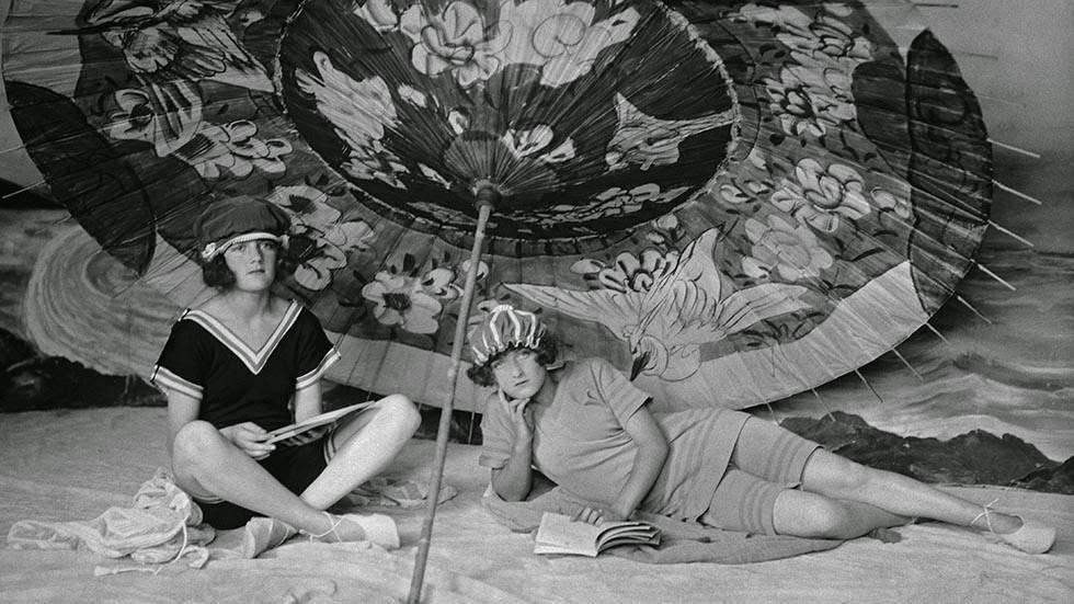 Two young women in swimming costumes reading under a large parasol, 1922