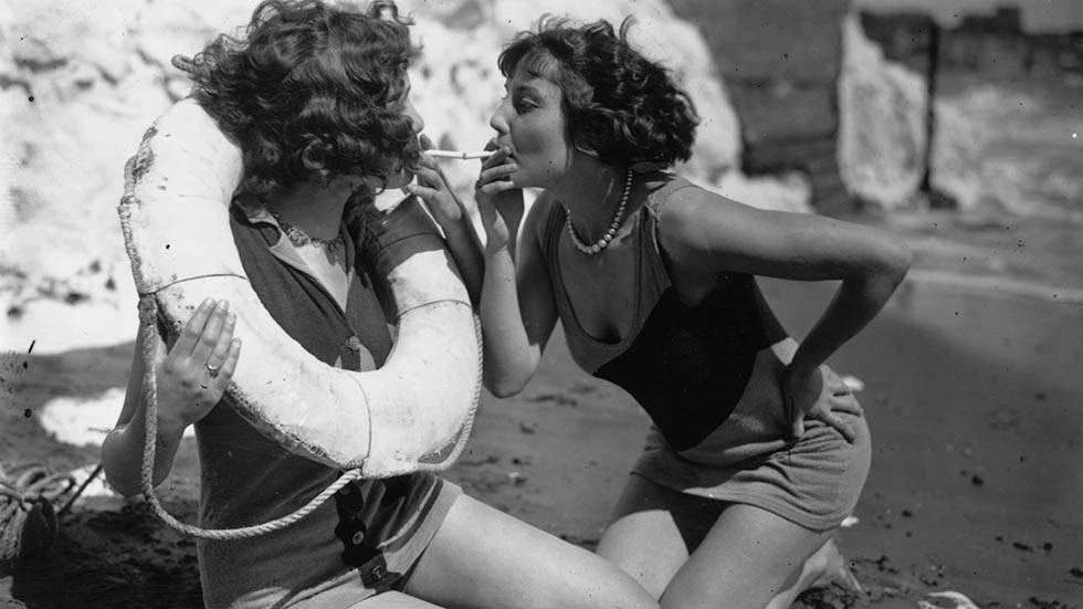Young bather lighting a cigarette with the tip of her friend's on the beach at Broadstairs, 1929