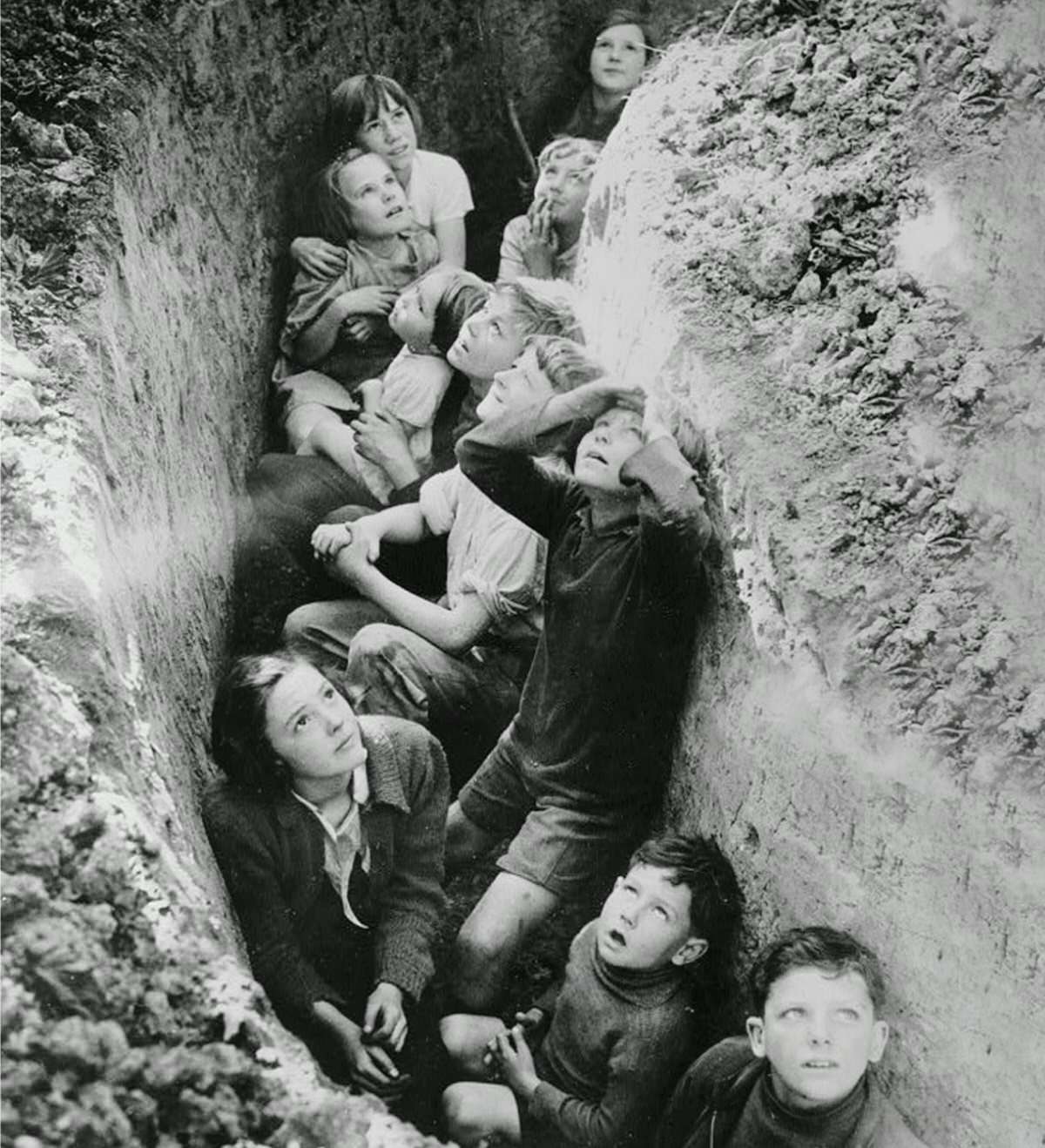 The children pictured above (circa 1940-1941) are crouching in a crude makeshift air raid shelter in the middle of World War II. Looks of shock and fear mingle with a kind of awe as they gaze towards the heavens. Many schoolchildren experienced close brushes with death during the war.