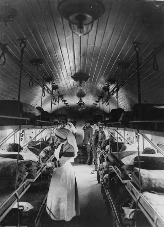 Interior of an ambulance-train ward, France, during World War I. This image is very striking due to the lighting and the tunnel effect of the train carriage, which is emphasised by the parallel lines of the wooden panelling on the roof. Two nurses are busy tending the wounded while two officers survey the scene from the top of the carriage.