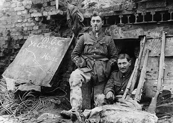 Some soldiers are jammed in the brick door way of a dugout while one man stands outside smoking. The two clearly visible soldiers look cocky and are wearing big grins. The dugout itself is half derelict with the structure clearly showing. The ground is very muddy and uneven. It is littered with equipment and debris. There is a sign leaning up against the wall which says 'ye corner house'.