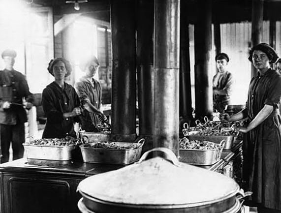 Women cooks at the Front, during World War I. Six flues from a group of Agas, which are arranged in a square in the middle of a wooden shed. On top of the Agas are big square metal tins with food piled over the edge of the dishes. The women all have implements sunk into the food like they are chopping it up. There are two soldiers standing in the kitchen with the women. One is uniformed and the other is dressed very informally. The four women are all shiny faced with their hair tucked up into 'mob- caps'. The are wearing big collared overalls on top of their clothes.