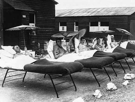 Five relatively young soldiers are lying in beds on a gravel path. The beds are made from pieces of metal tubing and look fairly compact. Each man has a number of pillows to prop him up and is holding a bamboo parasol. They are all sitting at different angles although it is unclear whether this is for medical reasons or just personal preference. The gravel path is marked with boulders which have been painted white. It skirts more wooden huts . The hut behind the soldiers has the number seven on it and a fire bucket is also visible.