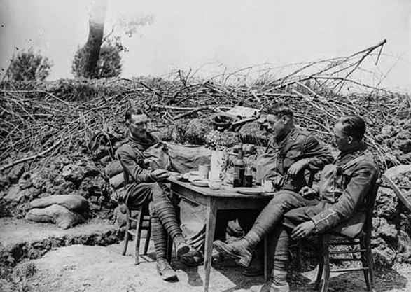Three officers of the Duke of Wellington's Regiment (West Riding) are sitting around a table. The table is laid with a vase of flowers, plates and mugs, but there is no food visible. The bottle appears to be labelled, 'Dark Port.' This photograph, which is attributed to John Warwick Brooke, shows the easier conditions often enjoyed by the officers.
