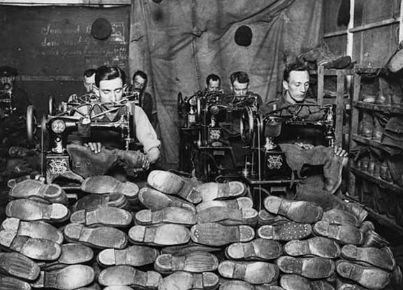This view of the corner of a workshop for repairing army boots shows men using machines to mend calf-length boots. The machines have the logo and brand name of the American Singer Company, who produced both domestic sewing machines and larger industrial machines such as these, for sewing leather and canvas.