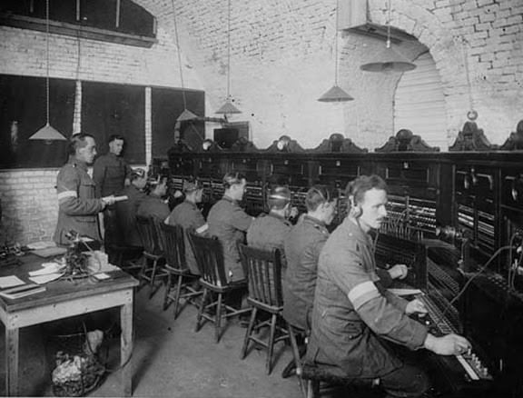 Signallers working at the headquarters of R.E.S.S. in France, during World War I. Just like a team of operators working at a busy telephone switchboard, this image shows the network of communications at company HQ that was required to co-ordinate an armys activities. The object immediately in front of the signaller on the right looks like a mouthpiece, which suggests that they are using wireless sets or field telephones to communicate.