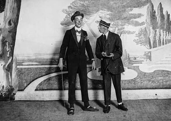 This image shows two entertainers on a stage, apparently in the middle of a stand-up comic turn. Both are in dark suits. One has a red clown's nose and a bowler hat. He is wearing dancing shoes, so probably did some sort of dance routine as part of the act. There is a painted landscape as a backdrop.