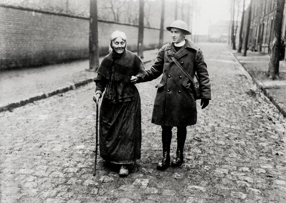 British priest helping a frail woman on a cobbled street in France, 1914