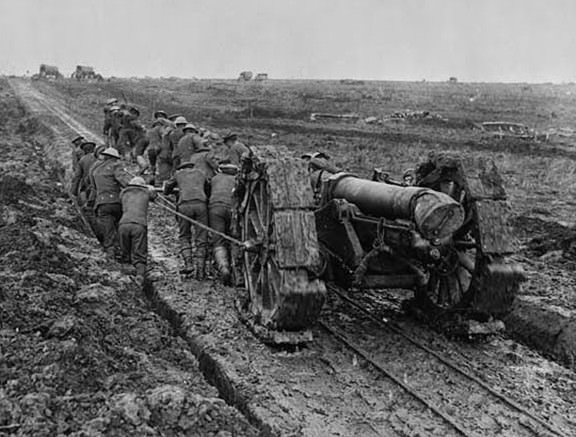 Soldiers struggling to pull a big gun through mud. The gun has been placed on a track created for a light railway. The soldiers are pushing a device, attached to the gun, that possibly slots into the tracks. Some of the men are in a ditch that runs alongside the track, the rest are on the track itself. A makeshift caterpillar tread has been fitted to the wheels of the gun, in an attempt to aid its movement through the mud. The surrounding landscape is bleak and desolate, with only a few trucks visible in the distance.