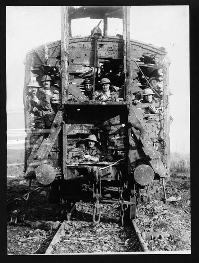Derelict train with soldiers standing in it, France, during World War I. The camera is face-on to the end of a blown out train carriage which is still sitting on its lines. The ground around the train is littered with branches and bark. The metal structure of the train is still intact but the wooden sides have little holes as well as big missing chunks. Seven soldiers are standing inside the carriage and looking out through these large gaps. They all appear to be quite relaxed and smiling.
