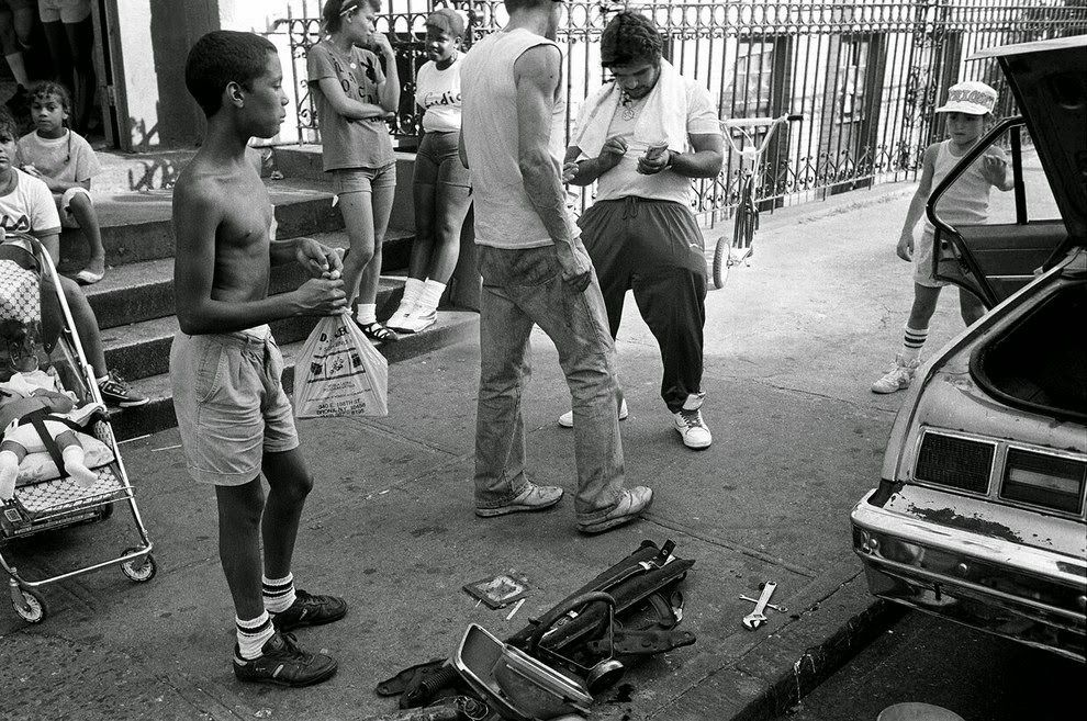 Dealer sells crack in front of a building as moms sit with their babies and children play, 1980s.