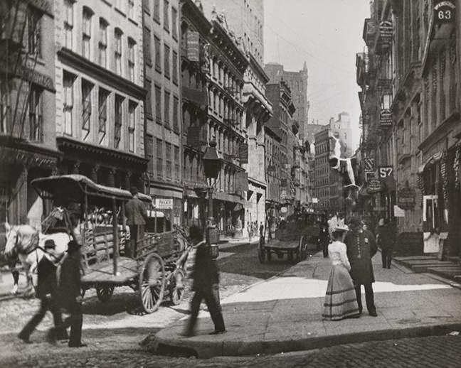 Woman talking to a police officer on Maiden Lane with delivery wagons in the street, 1891