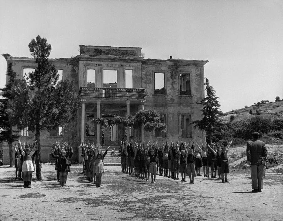 Children in a gymnastics class in front of a burnt-out school in Ghortiatis, Greece, 1948