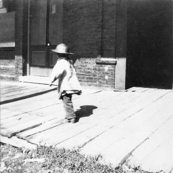 Child walking on wooden sidewalk in front of stores, 1897.