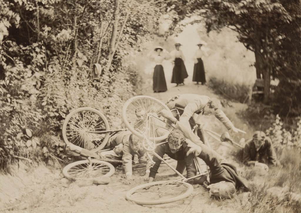 Four men and bicycles after a crash on a path with three women in the background, 1897