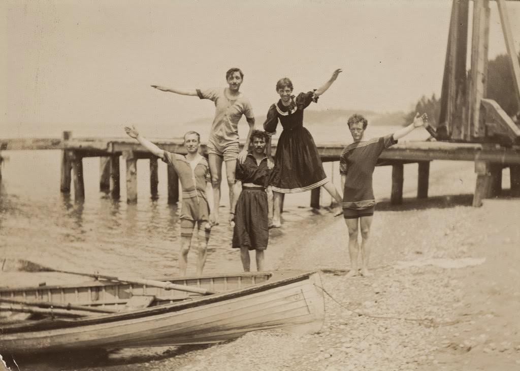 Five men, some in women's bathing attire, building a pyramid on a beach, 1897