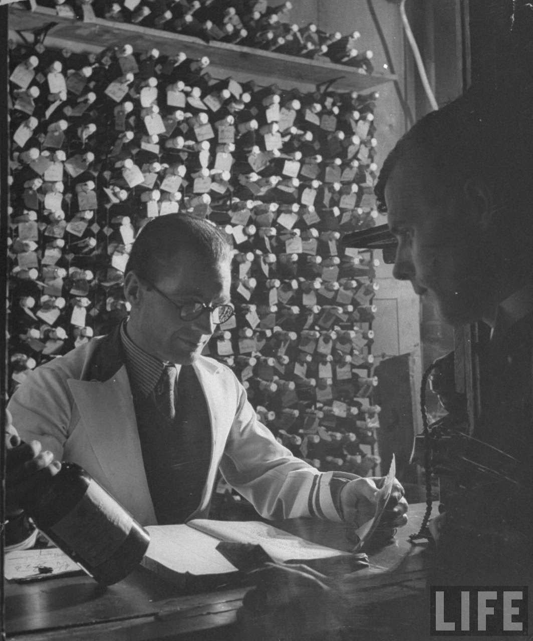 Barman checking the label on a member's bottle of liquor, 1940s