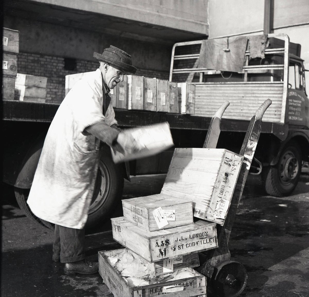 Elderly porter unloading wooden crates of cod fillet onto a trolley at Billingsgate Fish Market, 1950s