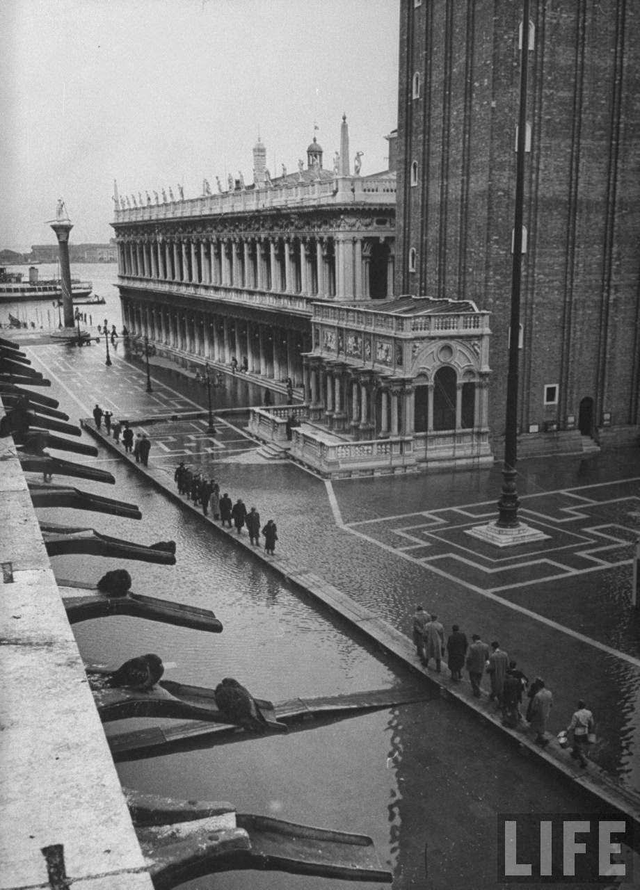 Pedestrians threading their way along makeshift wooden sidewalk across a flooded Piazza San Marco during its usual winter condition; Grand Canal is visible in distance.