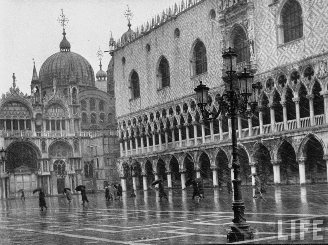 Pedestrians crossing flooded Piazza San Marco in front of the Basilica next to the Doge's Palace on a rainy day.