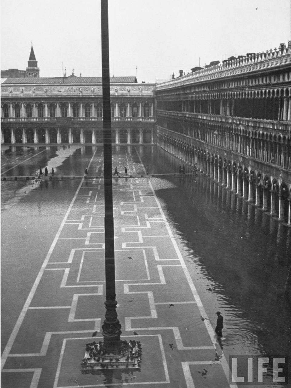 Pedestrians threading their way along makeshift wooden sidewalk across a flooded Piazza San Marco during its usual winter condition.