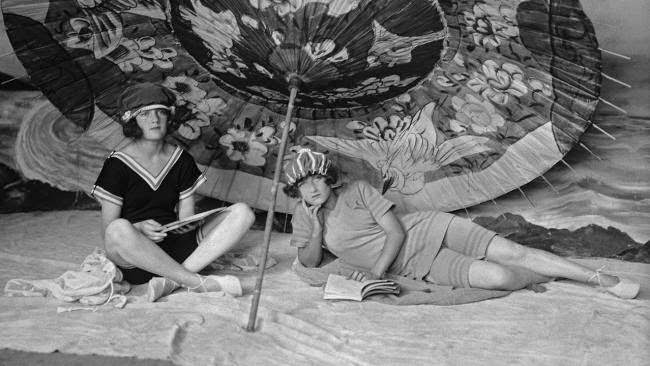 Two young women in swimming costumes reading under a large parasol, 1922