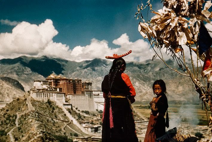 Mother and child pray on Chagpori's crest, a pilgrim shrine. Incense smolders beneath prayer flags.