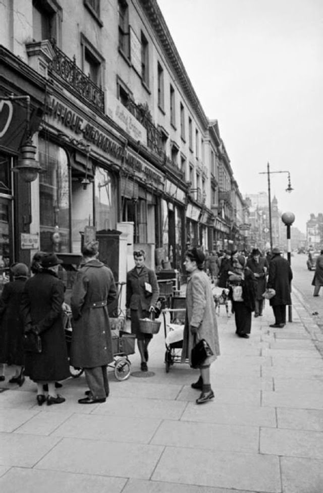After lunch, Mrs Day sets out to do her weekly shop on the King’s Road in Chelsea. She walks past several women with prams and a member of the RAF as they queue to the left of a large furniture store. The shop has furniture displayed on the street and the sign on its frontage says ‘Antique, Second-Hand and Modern Furniture’. Just above the heads of this group of people is a small sign directing people to a First Aid Post. In the background, other people go about their daily business and buses and cars are just visible in the distance. This photograph was almost certainly taken from a point on the King’s Road parallel to Walpole Road and is looking towards Sloane Square: the clocktower and spire just visible in the distance is on a building next door to Peter Jones department store.