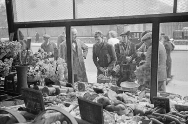 Mrs Day stops to look in the window of a shop to see what is available to her this week. The photograph is taken from inside the shop, and Mrs Day can be seen next to the shopkeeper. A group of other shoppers can also be seen. This photograph was probably taken on the King’s Road in Chelsea.