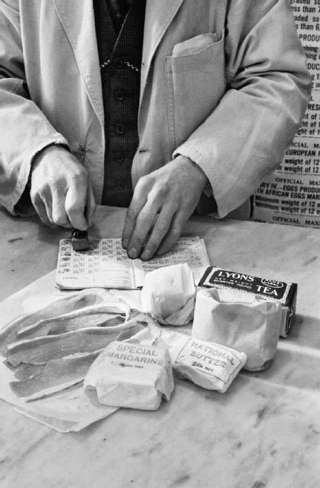 A shopkeeper stamps Mrs Day’s ration book during her shopping trip on the Kings Road in Chelsea. In the foreground can be seen the tea, sugar, ‘national butter’, margarine, cooking fats and bacon she is allowed for one week.