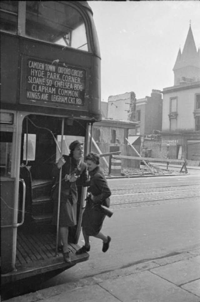 Mrs Day, helped by the female conductor, jumps on the bus that will take her to work. In the background, it is clear that quite a bit of air raid damage has been sustained. This photograph was probably taken on Fulham Road. The tower visible in the background is part of St Stephen’s Hospital (now the Chelsea and Westminster Hospital), which was built in 1878 as the Fulham Workhouse and St George’s Infirmary.
