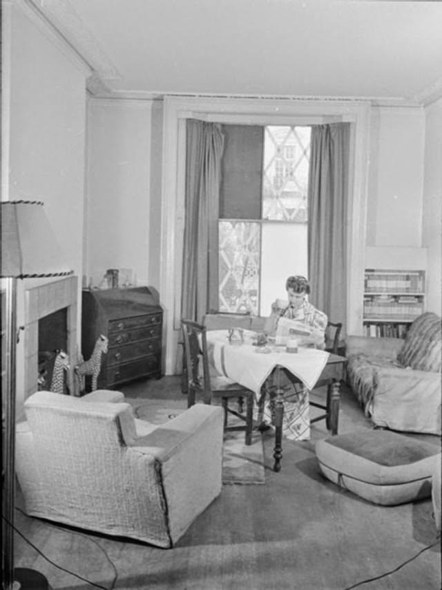 Mrs Day enjoys tea, toast and the morning papers at the breakfast table in the centre of her South Kensington sitting room. Behind her, evidence of air raids can be seen in that two panes of glass are missing from the window and have been replaced with boards and the other panes have criss-crosses of tape on them to prevent the glass from shattering, should the area suffer another air raid.