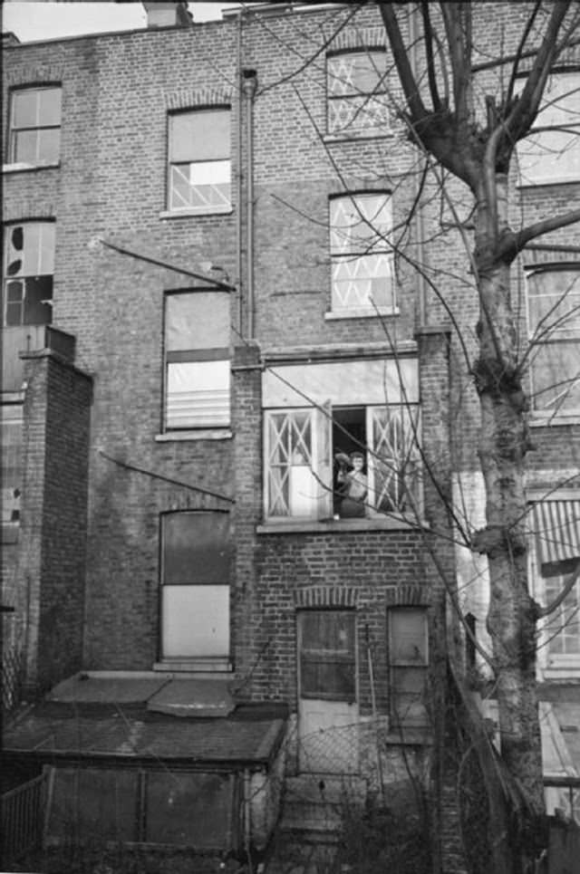 Mrs Day shakes her duster out of one of the back windows of her South Kensington home. Every visible window of her house, and of the houses alongside, bears witness to the air raids that have occured in the last few weeks. There is not one window that remains unaffected in someway and all are either fully or partly boarded, have had the broken glass replaced by oiled linen, or have existing glass criss-crossed with tape.