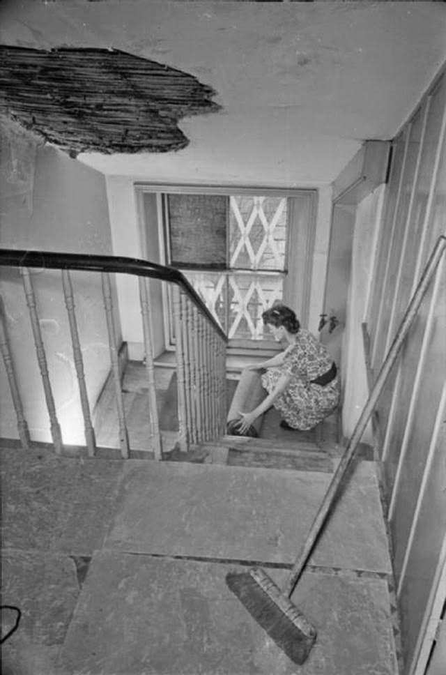 Mrs Day rolls away a rug that was on the staircase of her South Kensington home. All carpets have been removed and asbestos laid in their place, in an attempt to combat fire bombs. Behind her, part of the window has been boarded up, with the rest of the panes have criss-crosses of tape across the glass.