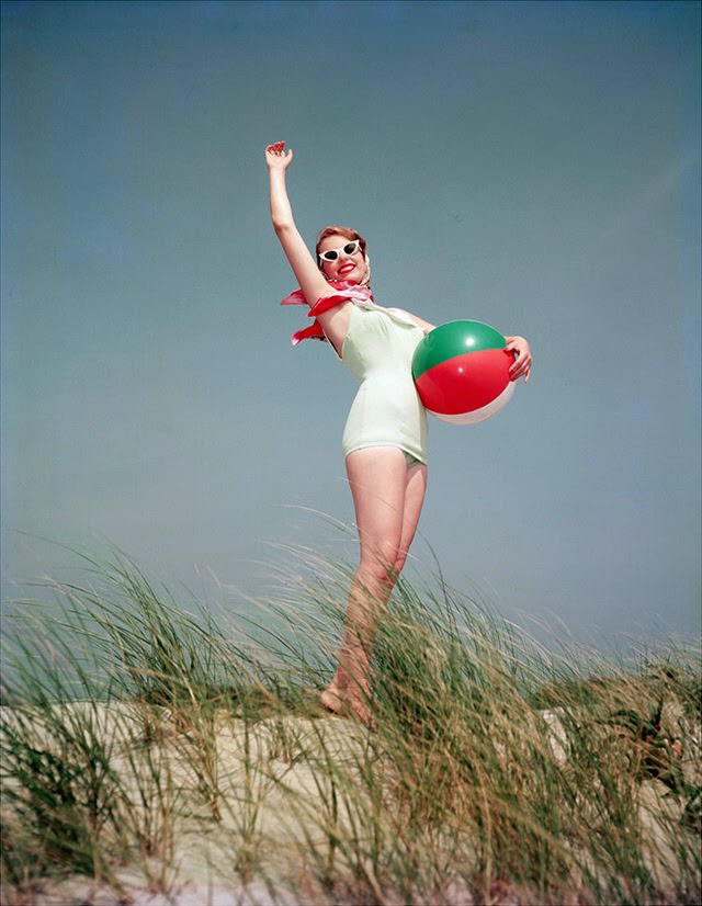 Young woman in a bathing suit holding a beach ball on a sand dune, 1956