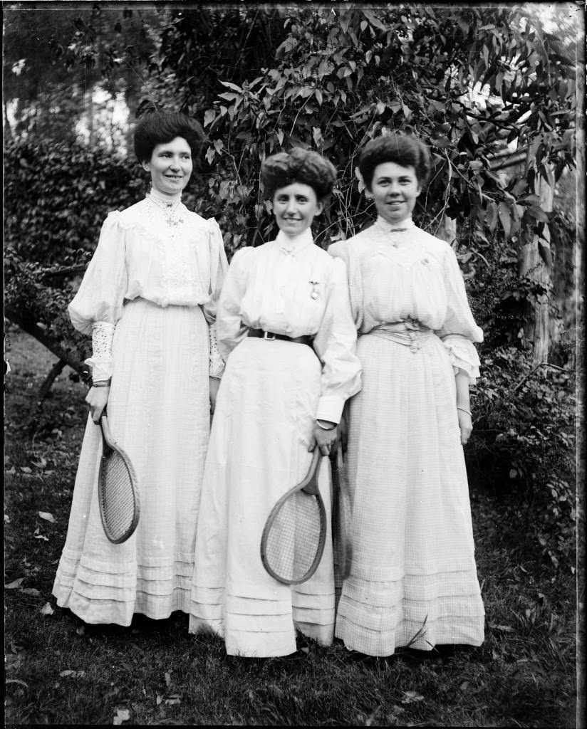 Three young women in light dresses holding tennis racquets, 1900