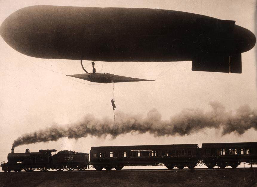Zeppelin flying low over a steam locomotive with a man on a rope.
