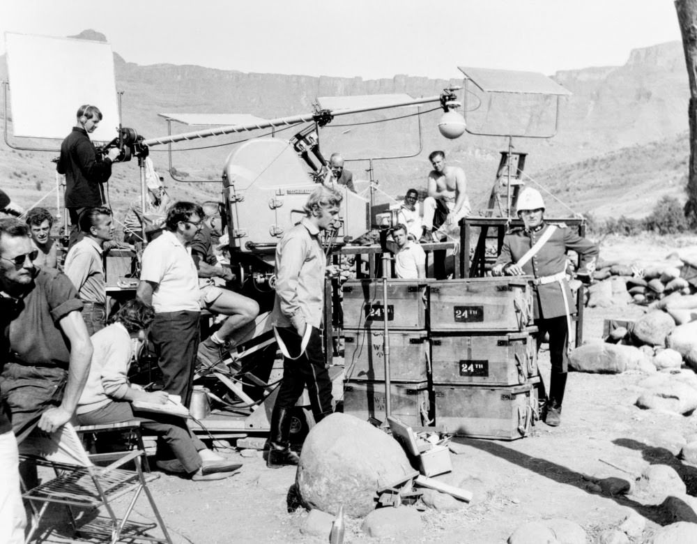 Michael Caine enjoys a moment out of full costume, as the lighting crew prepare the next shot on location for Zulu (1964). Top-billed Stanley Baker (right), meanwhile, is top-to-toe in helmet and bright red uniform.