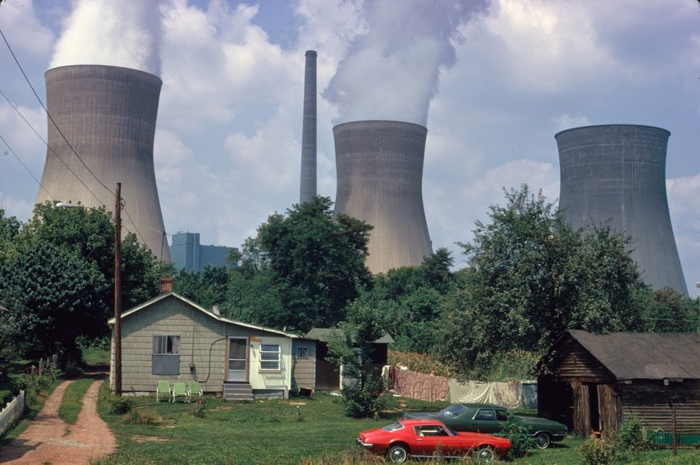 Water cooling towers of the John Amos Power Plant loom over a Poca, West Virginia, home that is on the other side of the Kanawha River. Two of the towers emit great clouds of steam, 1973