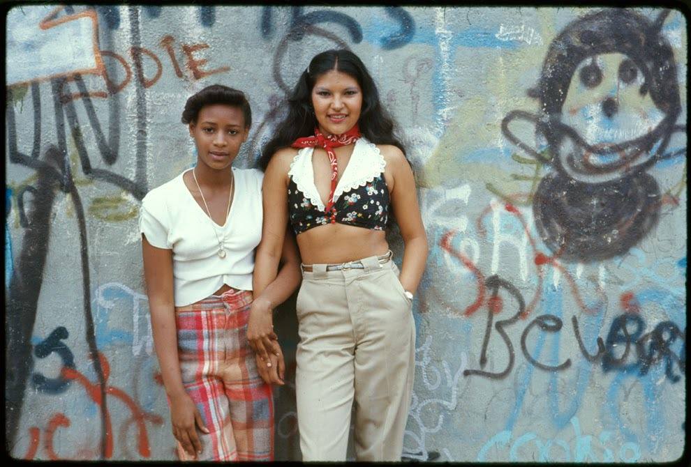 Two Latin girls pose in front of a wall of graffiti in Lynch Park, Brooklyn, New York, 1974