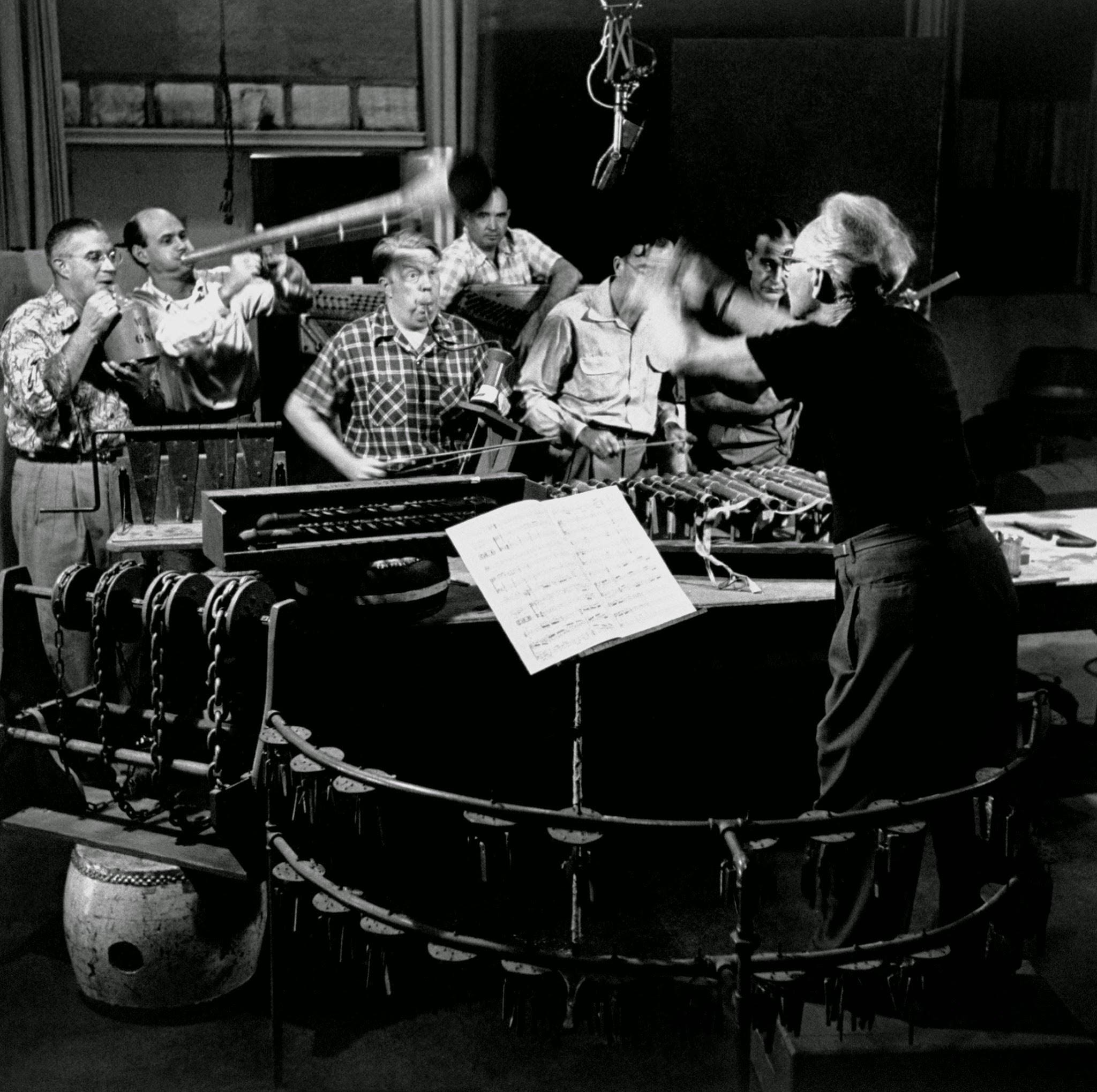 Jim MacDonald, Disney's sound effects expert, blows across top of jug while other experts make murderous music out of bazooka, disembodied piano and assortment of strings, pipes, hinges, chains, 1950s