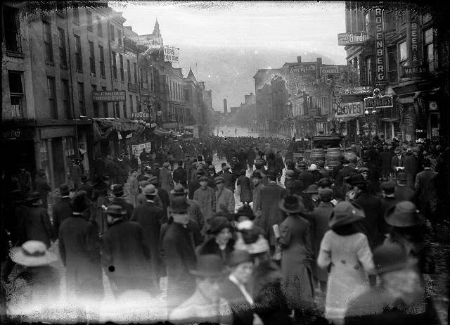 A large crowd is gathered on Front Street to look at the flood at the other end of the street. Varlan's Hotel, Rosenberg's Pawnshop, Charles Adam's Sons Grinding, J.G. Zweigle's Sons, W.T. Bridle Furniture, William Pigeon Shoes, Hyman Davis Shoes, Genesee Provision Company, Myers Department Store, and the F.H. Loeffler Company are visible, 1913
