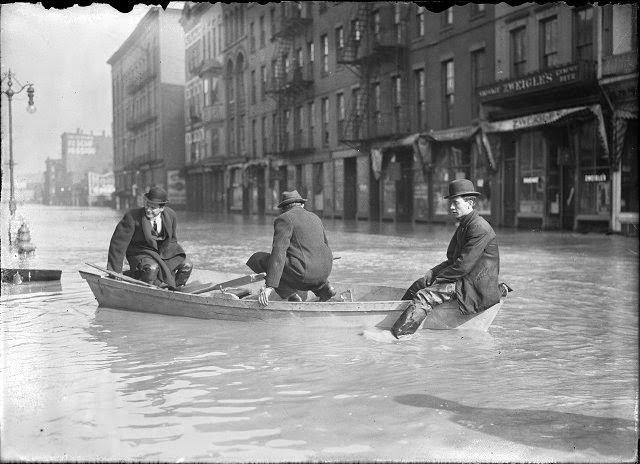 Three men ride a rowboat down flooded Front Street. They are passing J.G. Zweigle's, 1913