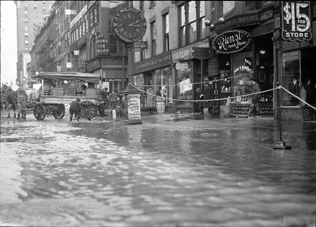 The flood has filled the store cellars with water on Main Street East at Graves Street. All the first floors are submerged as well. A horse and wagon are at the curb and the men are standing on the sidewalk. F.P. Van Hoesen Company, Rienzi (a bar), J.C. Wilson Company, the Postal Telegraph Cable Company, and the Fifth Avenue Clothes Shop are visible, 1913