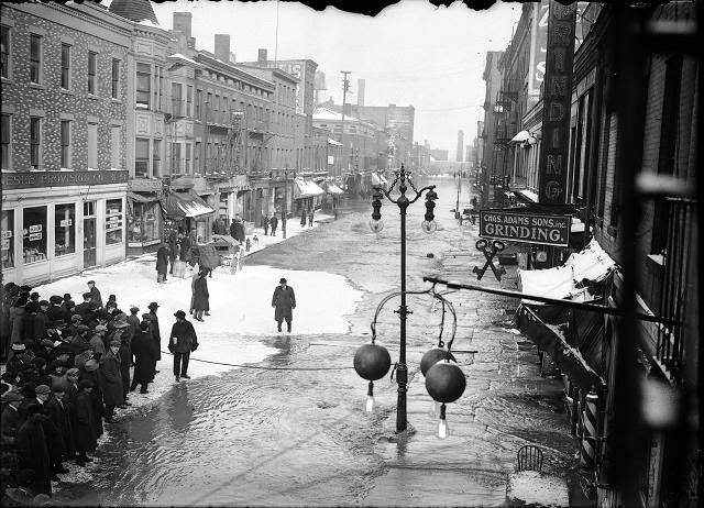 Rochesterians viewing the water on Front Street. Ice has formed in pockets on the flooded street. The sign for Charles Adam's Sons, Inc., 1913